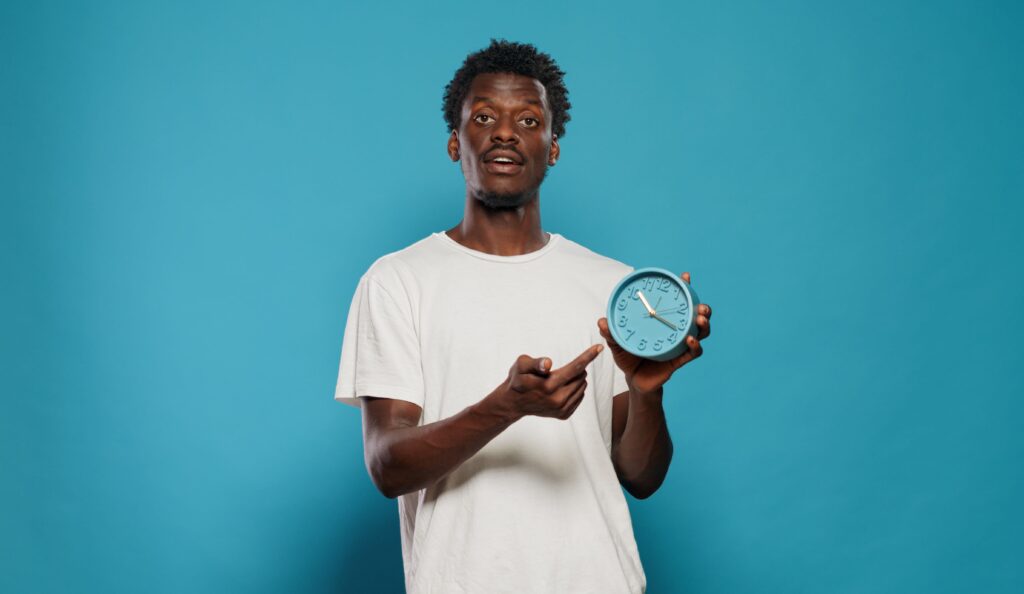 A man holding a blue alarm clock and pointing at the time