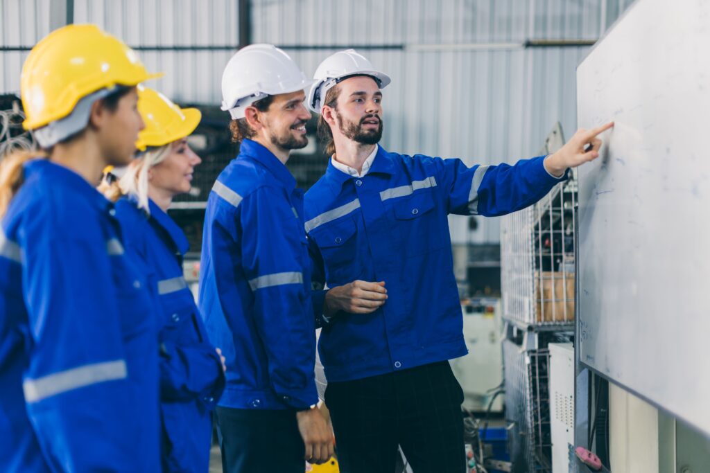 Blue collar workers huddled together listening while a man points at a whiteboard